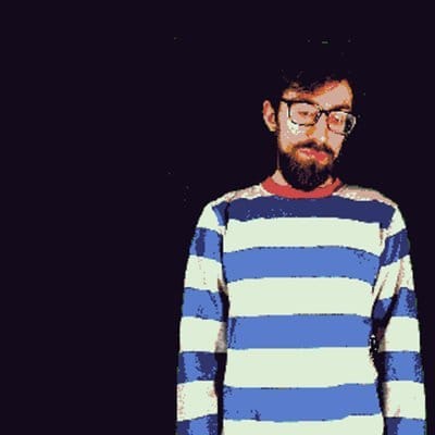 A portrait of SEQUENCEBREAK// curator Nilson Carroll. He wears a blue and white striped shirt and glasses. He has dark hair, a beard and a contemplative look on his face.